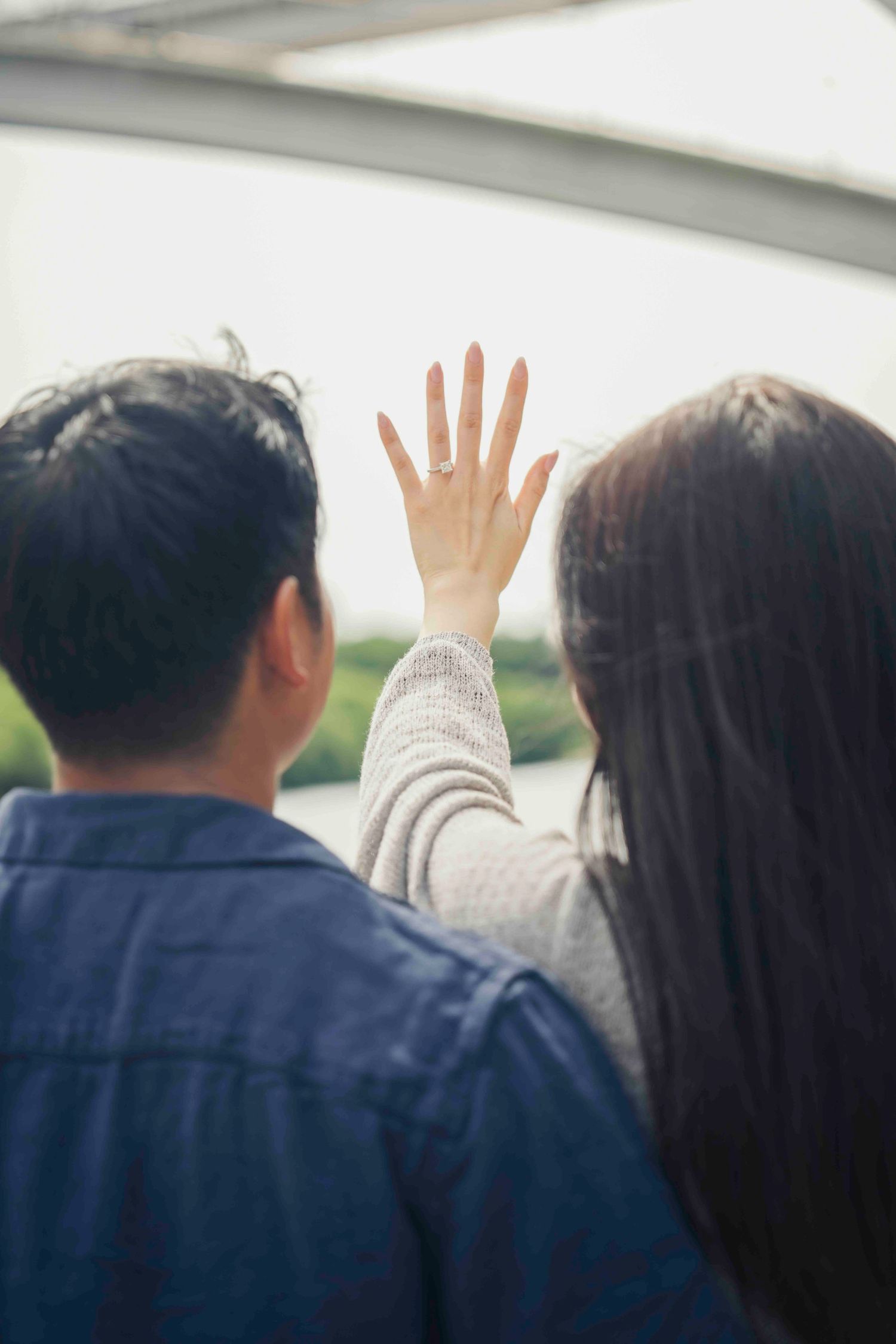 Couple viewed from behind, woman raising hand showing engagement ring, standing together near bright window.