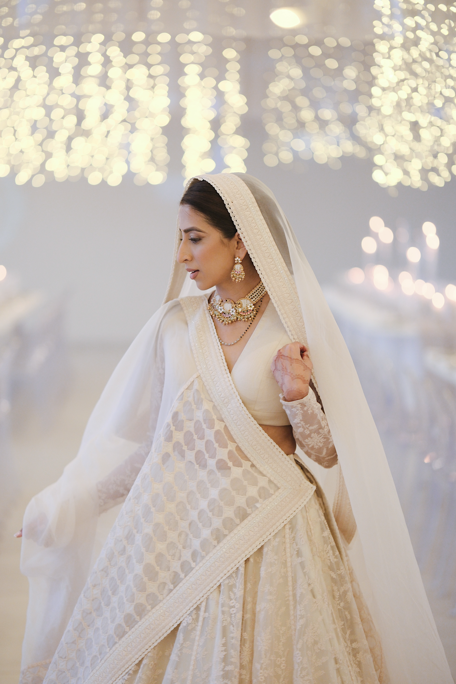 Indian bride in ivory lehenga with dupatta and gold jewelry poses under sparkling chandelier.