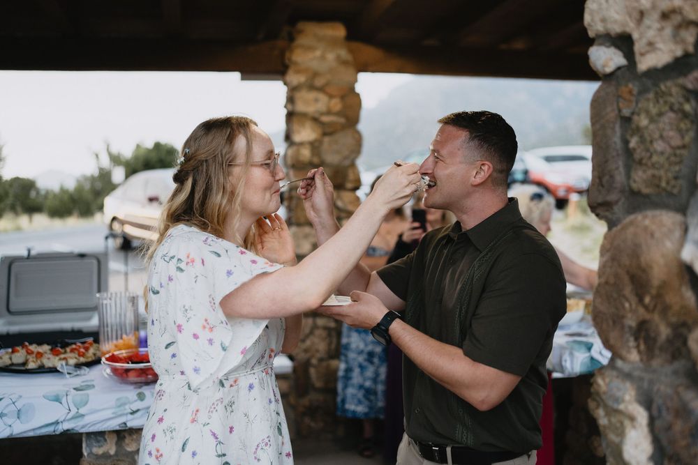 Couple playfully feeding each other at an outdoor wedding reception under a stone pavilion.