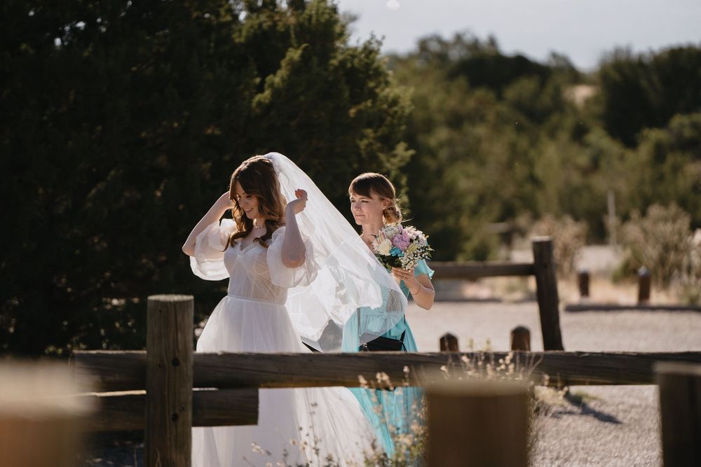Bride adjusting her veil with a bridesmaid holding bouquet near a wooden fence outdoors.
