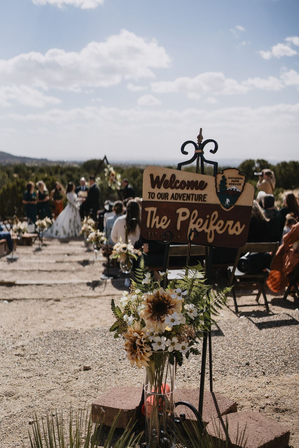 Rustic outdoor wedding aisle with a wooden The Pfeifers welcome sign and wildflower arrangements in Elena Gallegos Trailhead