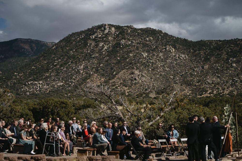 Wedding guests seated outdoors during ceremony with dramatic mountain landscape and cloudy sky in background at Elena Gallegos Trailhead