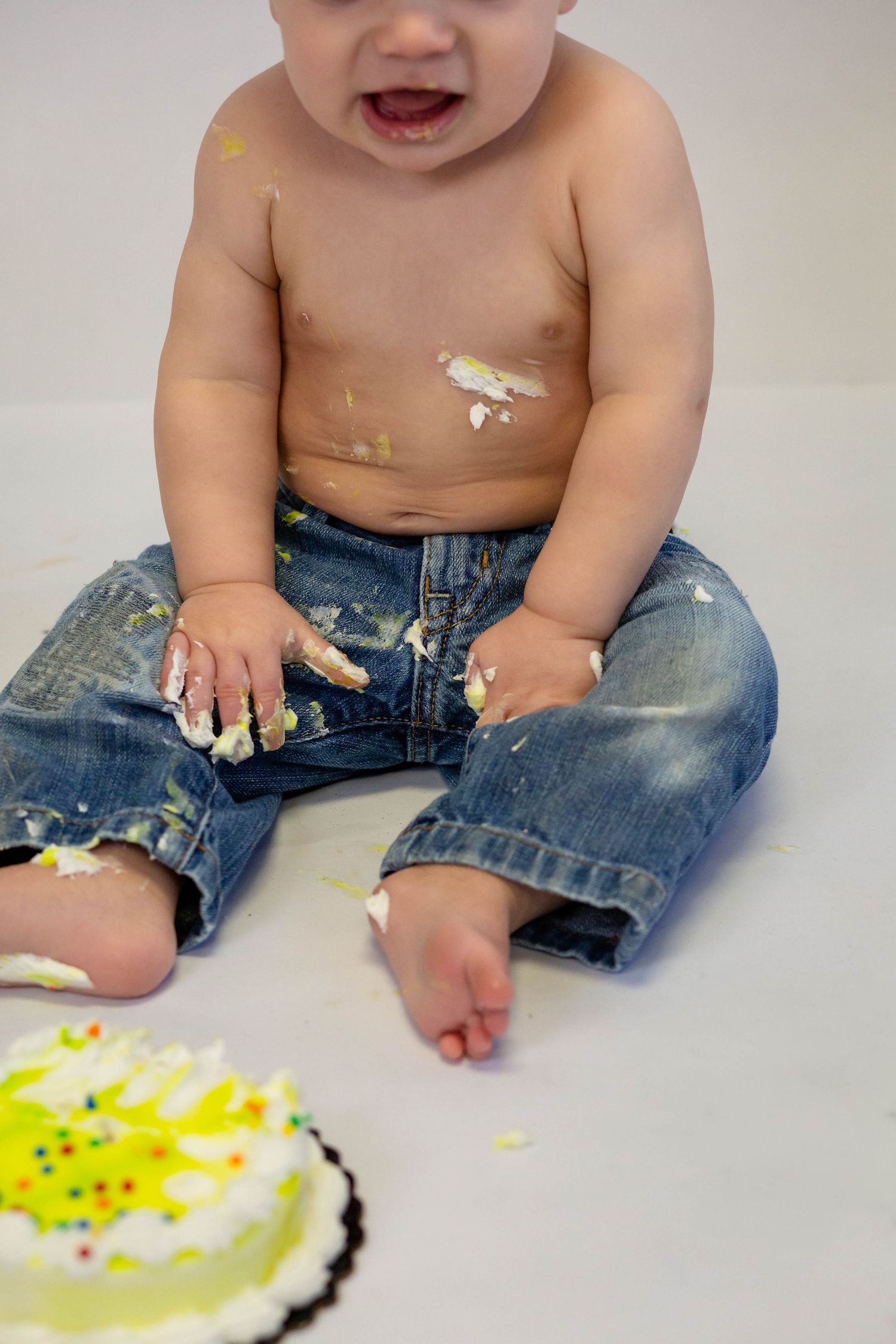 Shirtless baby in jeans smashing a white birthday cake, covered in frosting, celebrating first birthday.