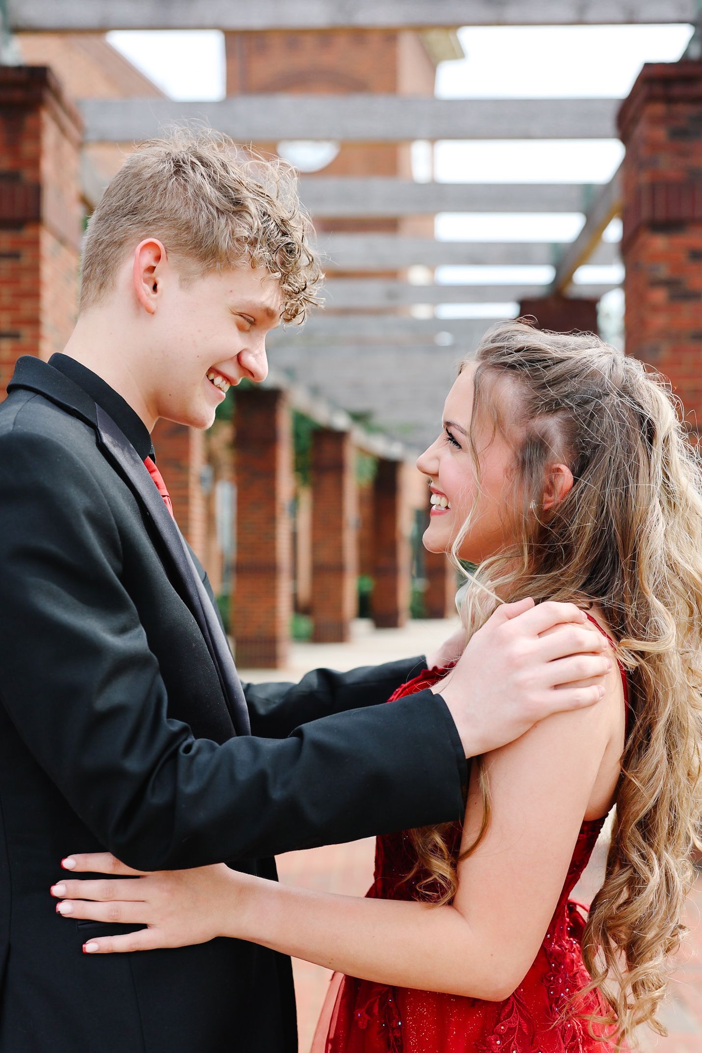 A smiling teenage couple in formal prom attire embrace outdoors, the boy in a black suit and girl in a red dress.