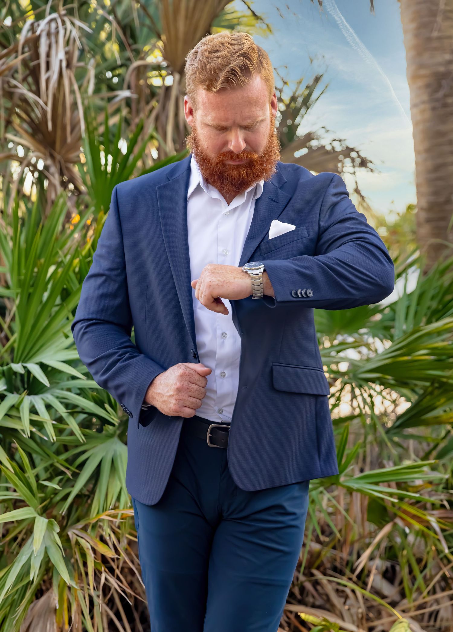 A red-bearded man in a navy blue suit adjusts his sleeve outdoors among tropical palm trees.