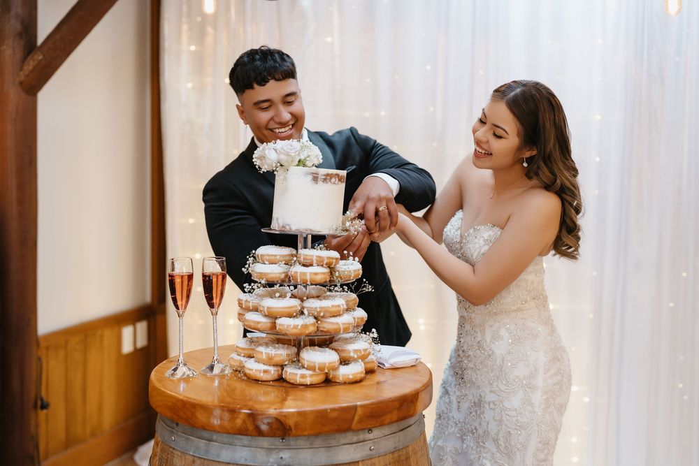 Bride and groom cutting a tiered donut wedding cake tower on a barrel, with champagne glasses nearby.