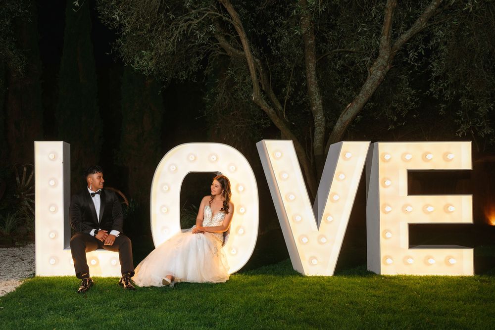 Bride and groom sitting together beside illuminated LOVE marquee letters on grass at an outdoor evening wedding.