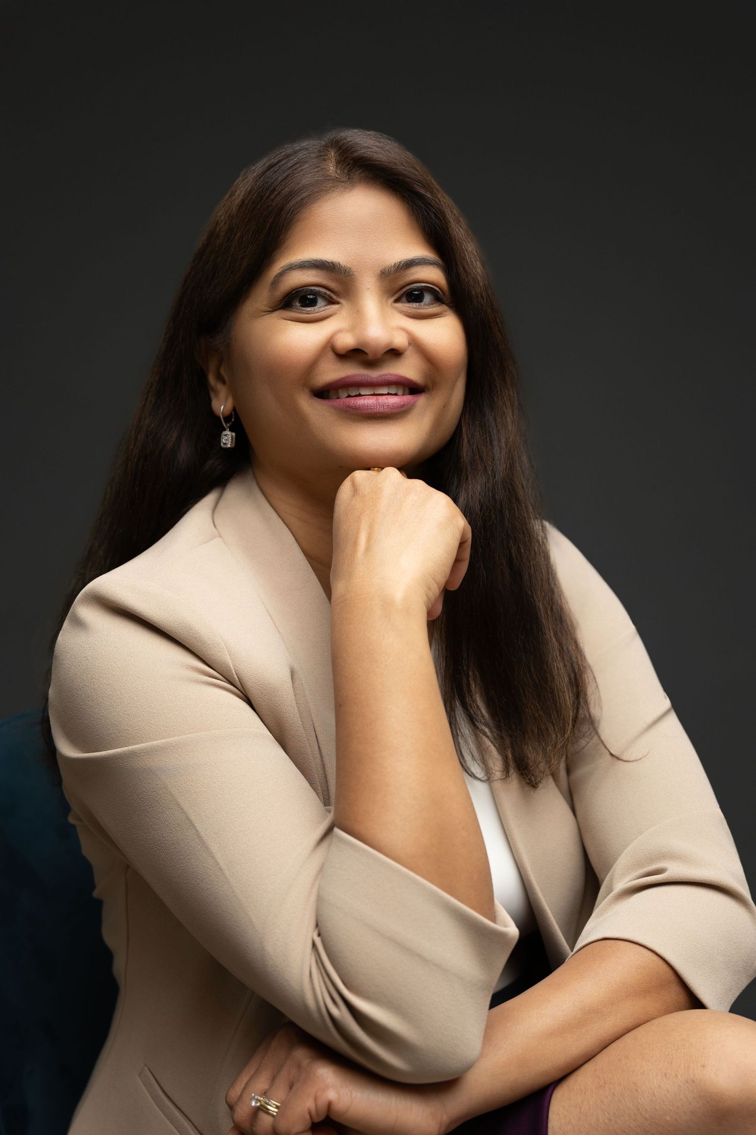 Professional woman in beige blazer smiling confidently with hand on chin against dark background.