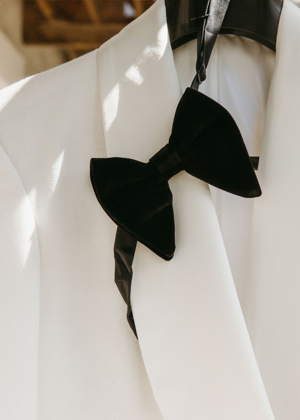 A black velvet bow tie hanging on a white dress shirt on a hanger in soft natural light.