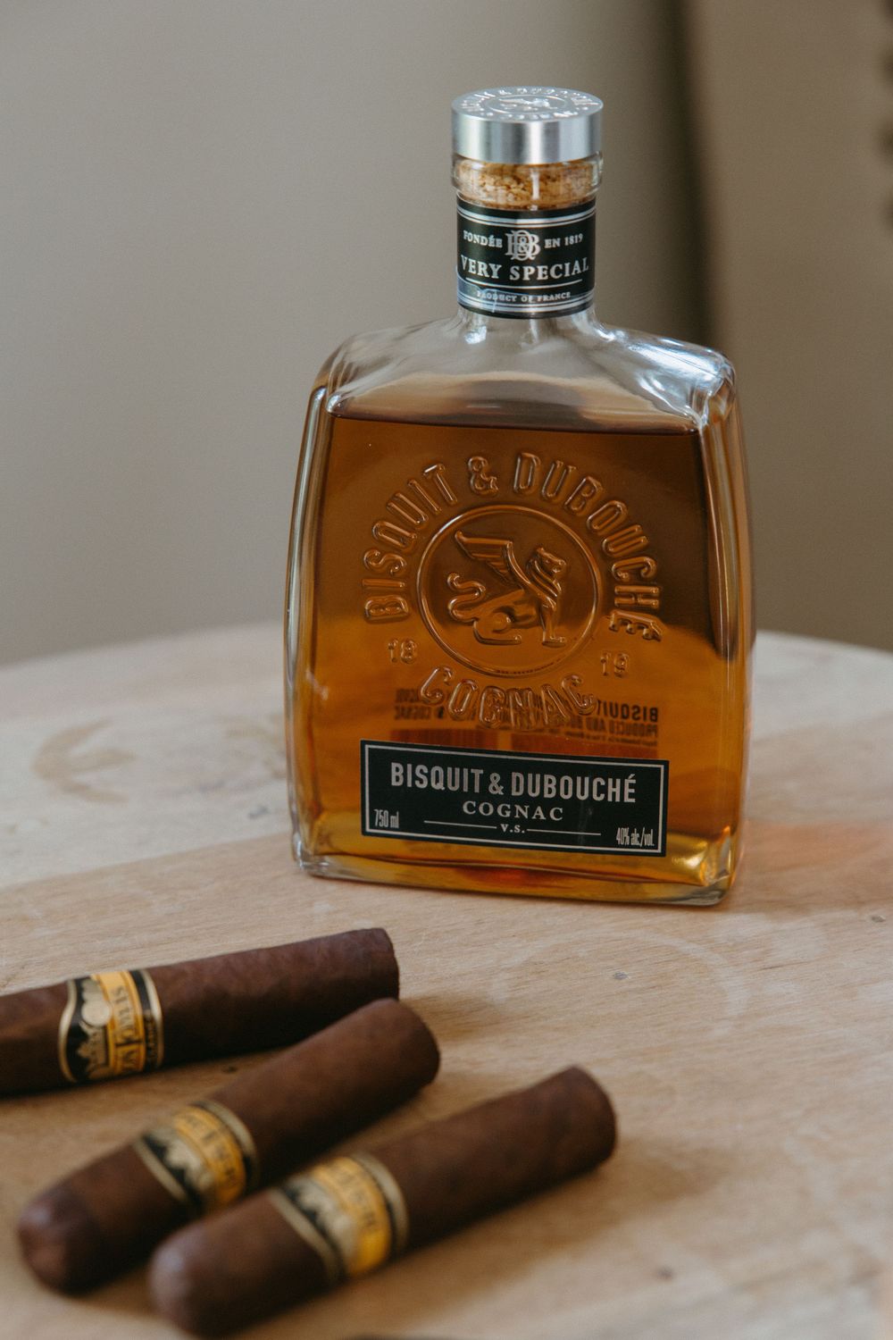 Bisquit & Dubouché VS Cognac bottle beside three cigars on a wooden surface.