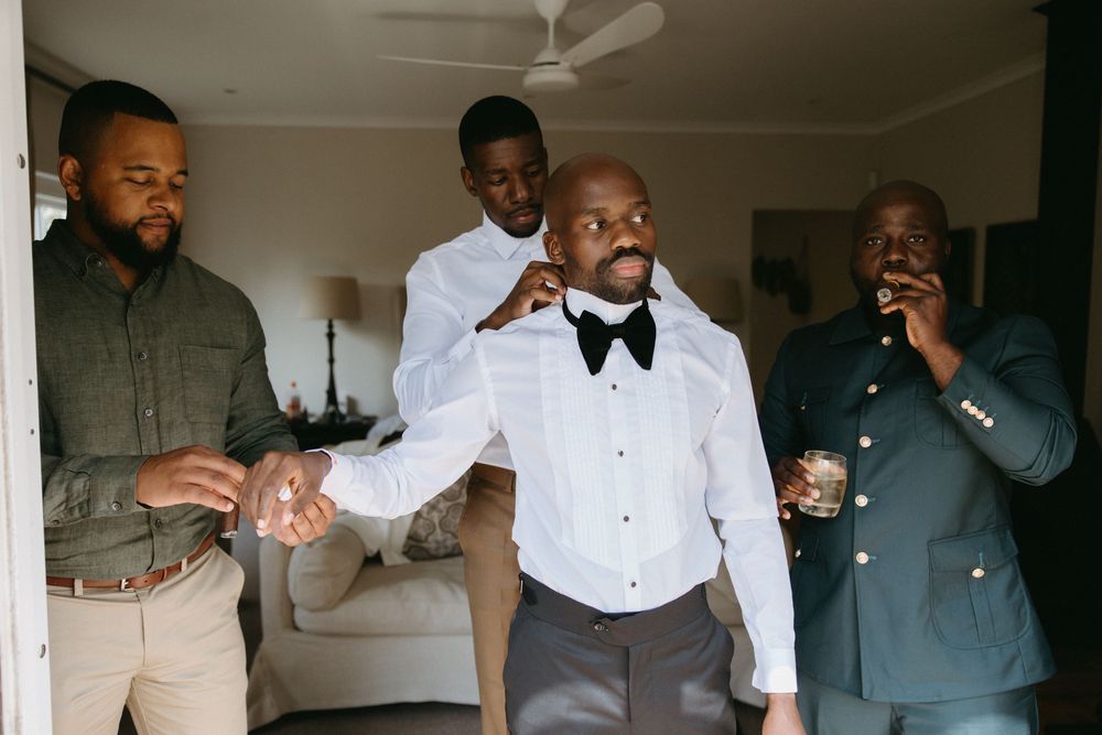 Groomsmen helping groom adjust bow tie and suit before wedding ceremony indoors.