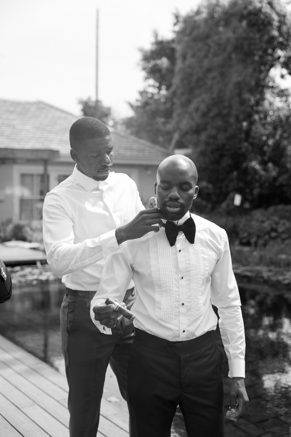 Best man adjusting groom's bow tie outdoors before wedding ceremony in black and white photo.