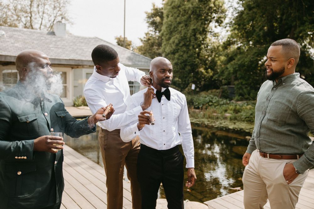 Groom and groomsmen socializing outdoors near a pond, one smoking a cigar, during wedding preparations.