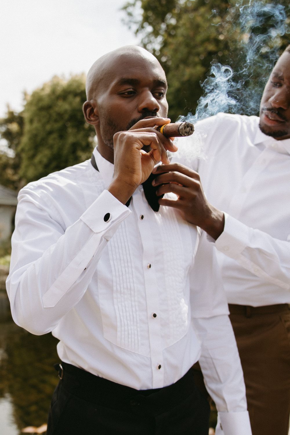 A bald Black man in a white tuxedo shirt and bow tie smokes a cigar outdoors while another man assists him.