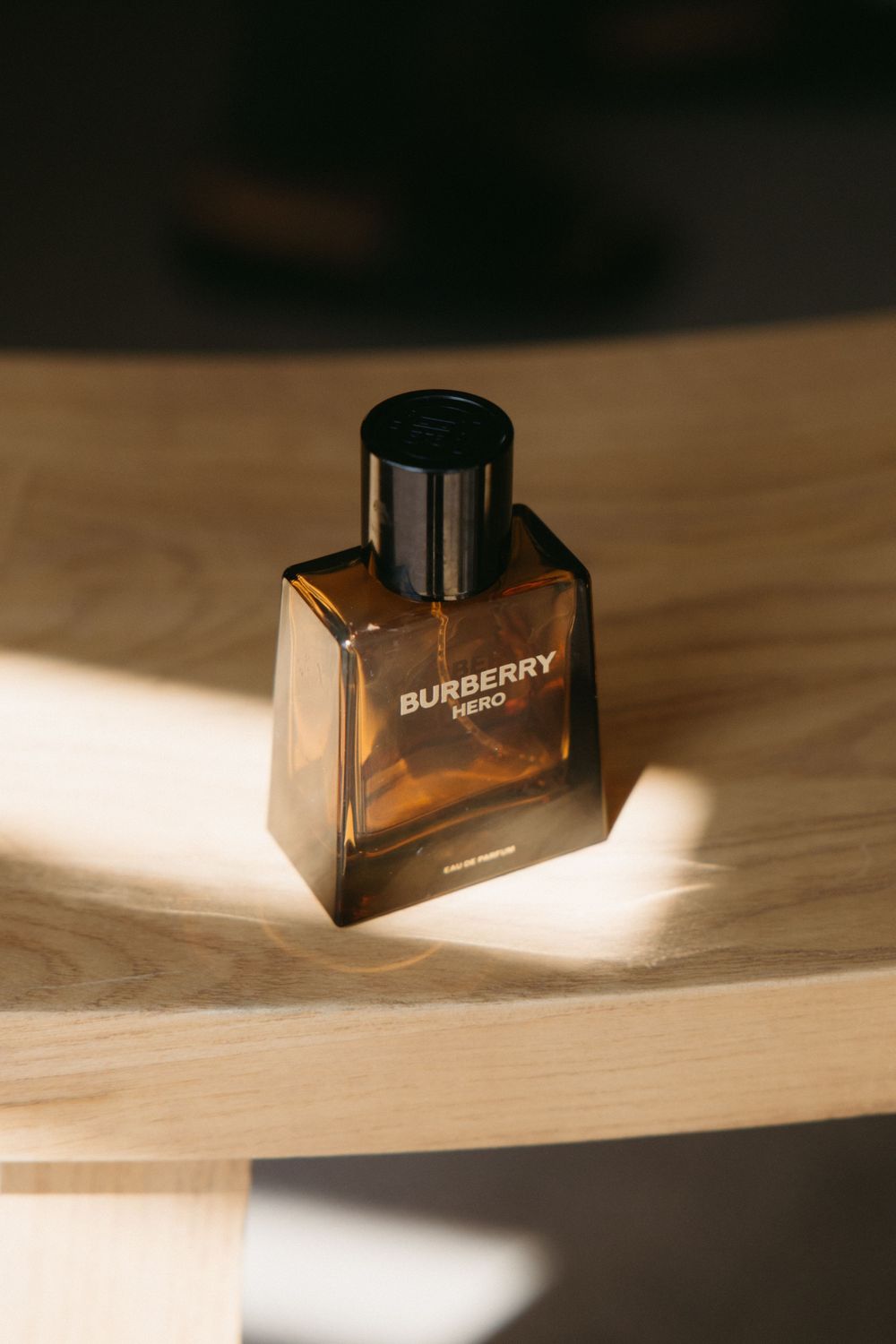 Burberry Hero Eau de Parfum bottle in amber glass with black cap on wooden surface in warm sunlight.