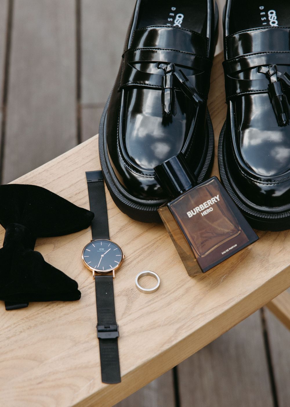 Black patent leather loafers styled with a watch, ring, bow tie, and Burberry cologne on a wooden table.