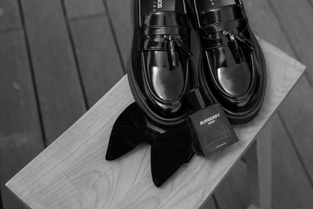 Black leather tassel loafers beside a velvet bow tie and Burberry cologne on a wooden surface.