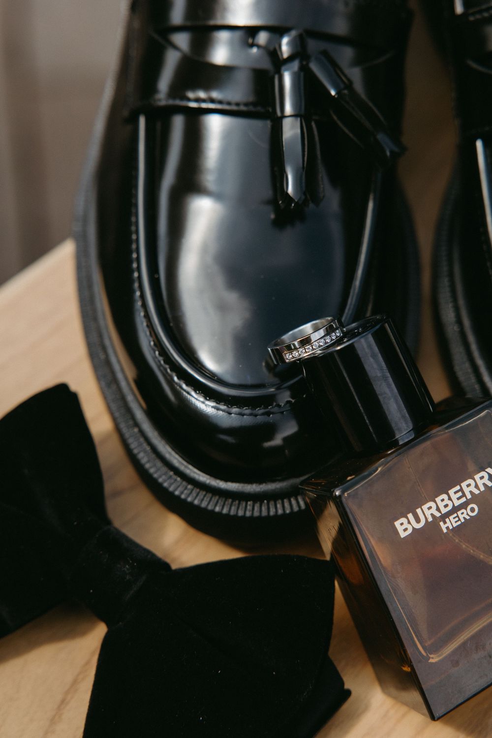Black tassel loafers, Burberry Hero cologne, wedding ring, and black velvet bow tie arranged on a wooden surface.