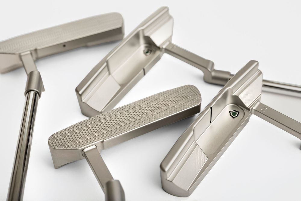 Multiple silver golf putters arranged on a white background, showcasing various blade and mallet designs with textured faces.