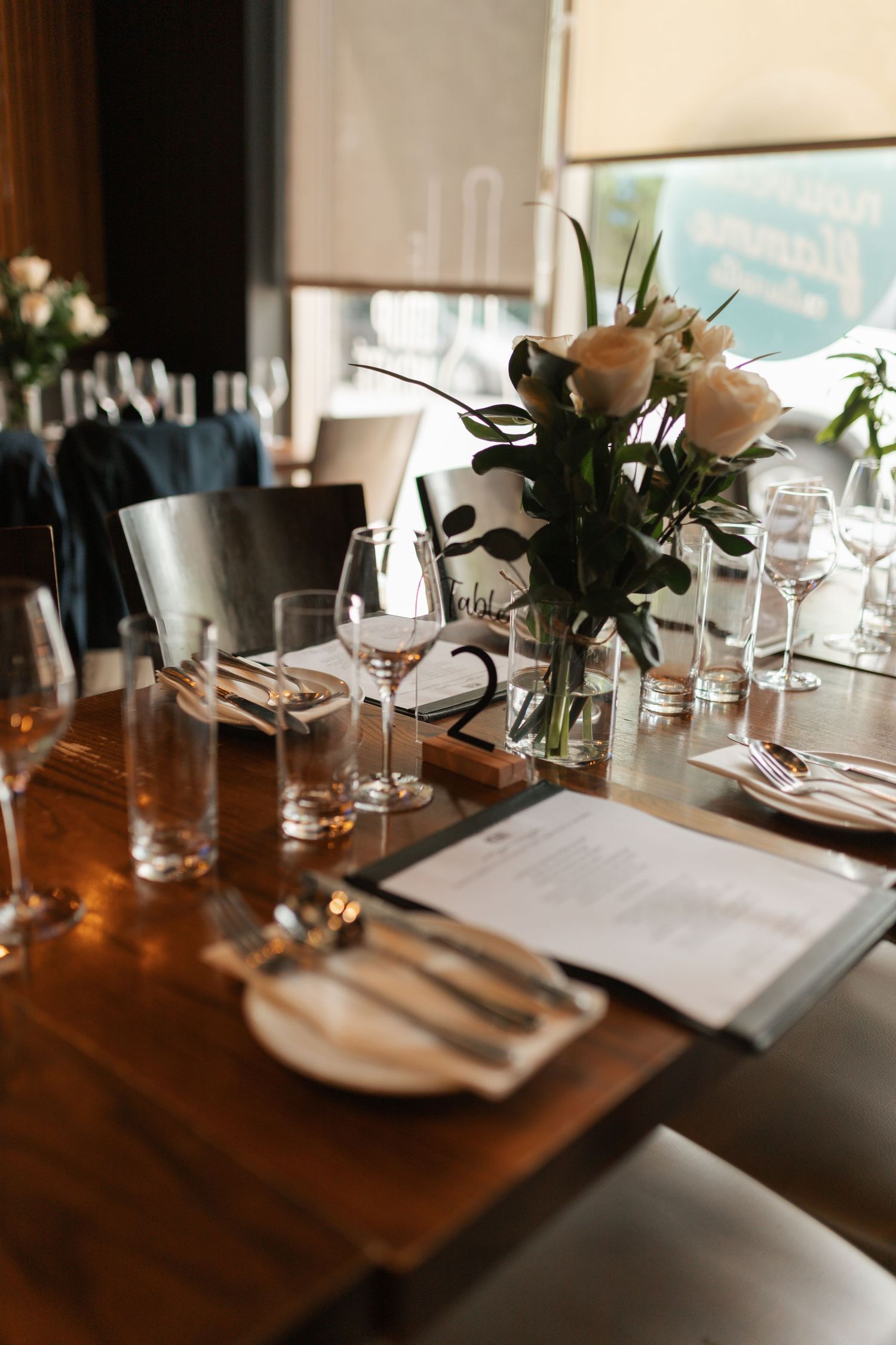 An elegantly set wooden dining table with roses, wine glasses, menus, and cutlery at a wedding reception venue.