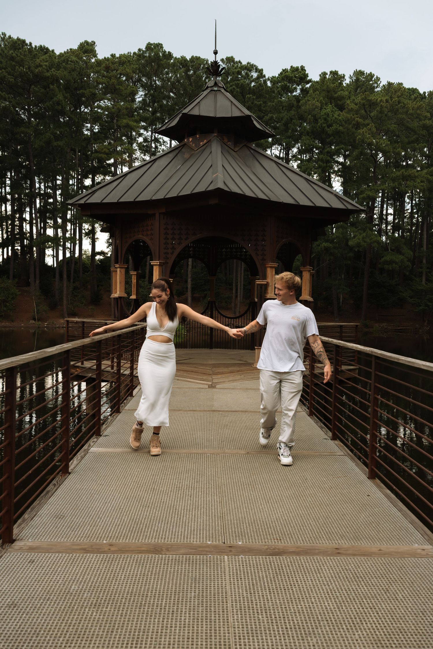 Couple holding hands walking on a bridge toward a brick gazebo surrounded by trees and water.