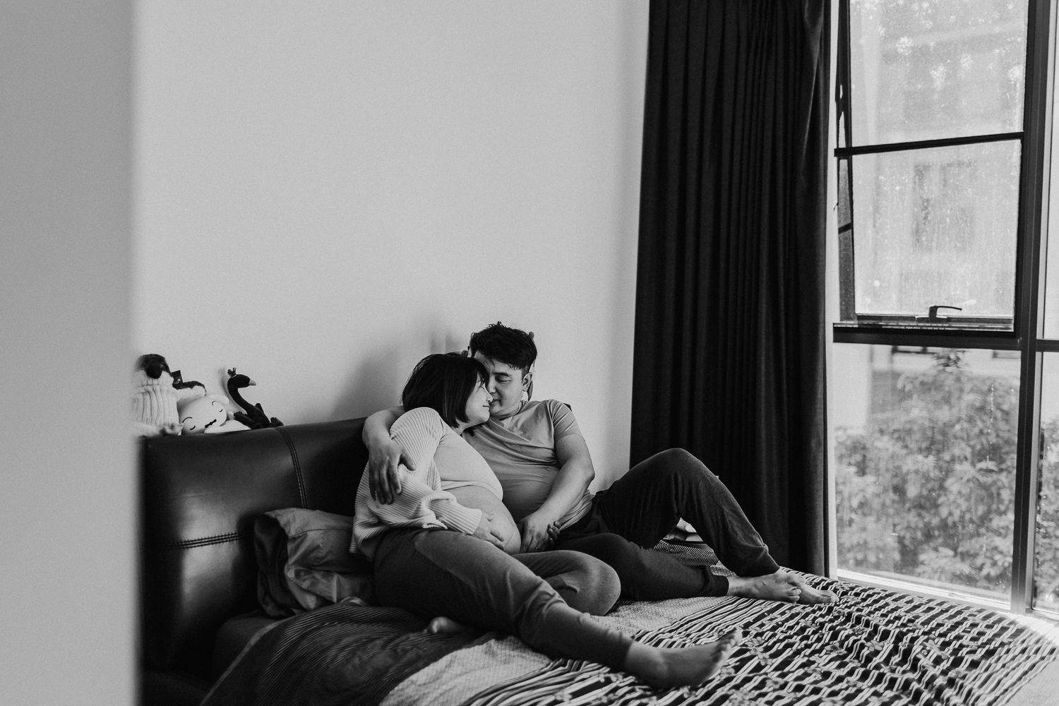 Black and white photo of a couple sitting intimately together on a bed near a window.