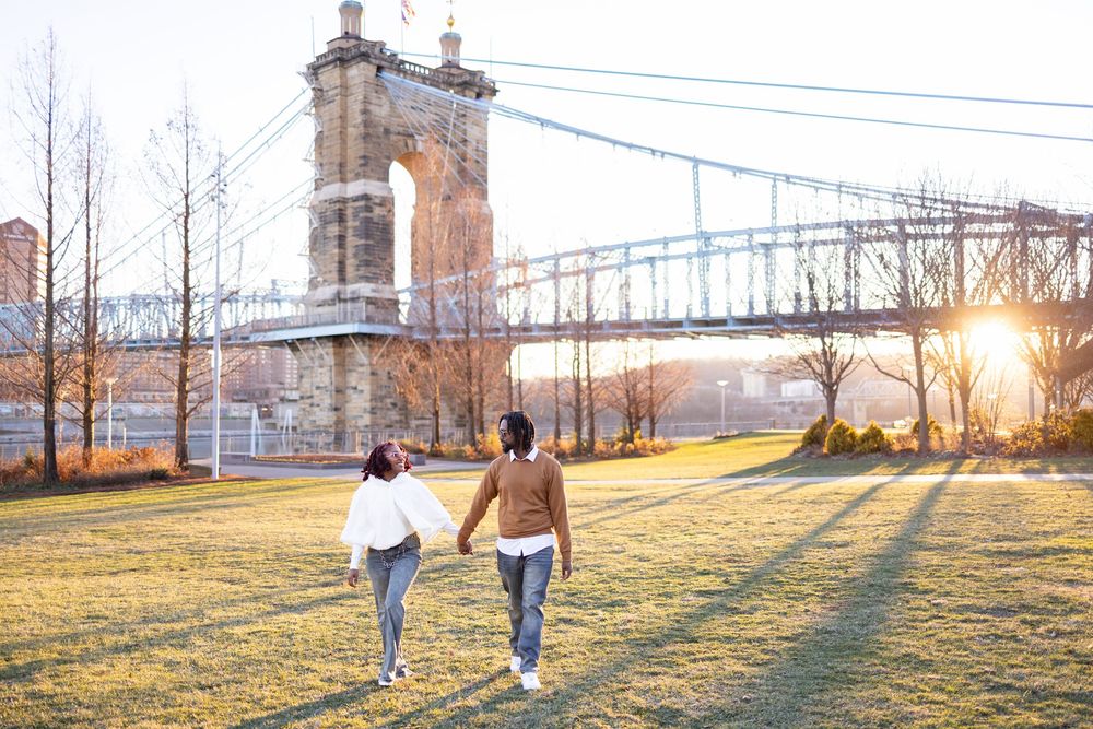 Couple holding hands walking toward a suspension bridge at golden hour sunset in Smale Park during their fall engagement photoshoot.