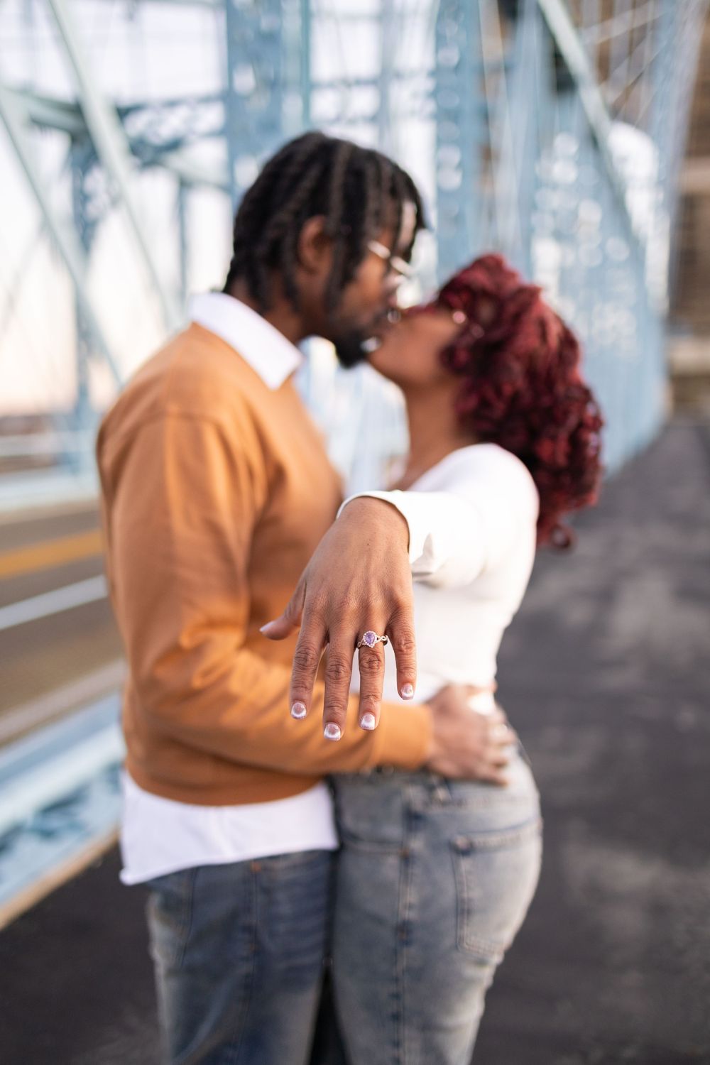 Couple kissing on a bridge while showing off an engagement ring, celebrating their proposal outdoors.