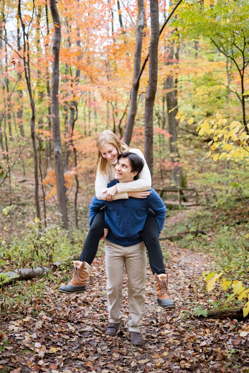 Couple enjoying a piggyback ride on a trail surrounded by colorful autumn foliage in Ault Park during the fall cincinnati engagement shoot.
