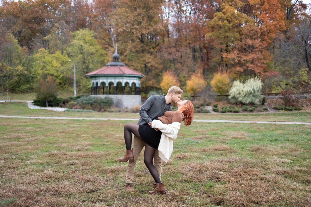 Couple dancing romantically in autumn park with colorful foliage and gazebo in background.