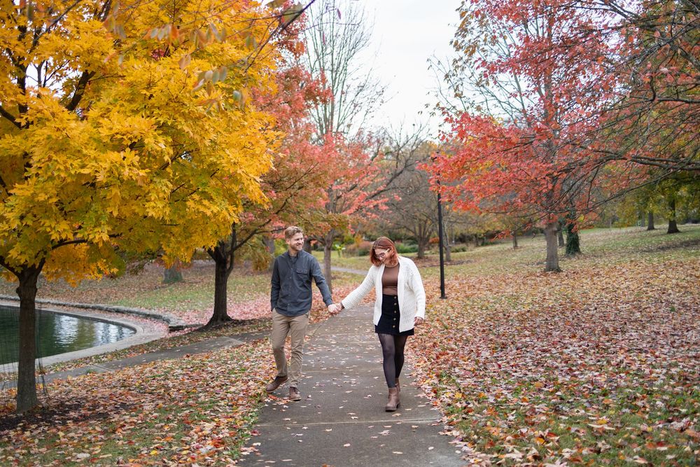 Couple holding hands walking on a leaf-covered path surrounded by vibrant autumn foliage in scenic Eden Park in Cincinnati.