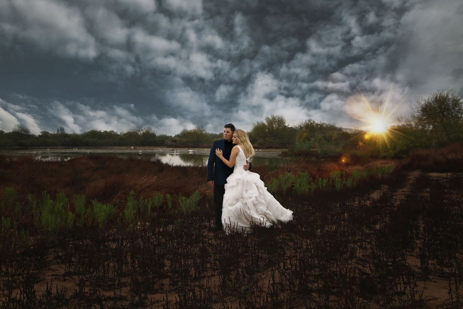 Bride in a white gown and groom in a dark suit embrace in a marsh field beneath dramatic storm clouds and a glowing sunset.