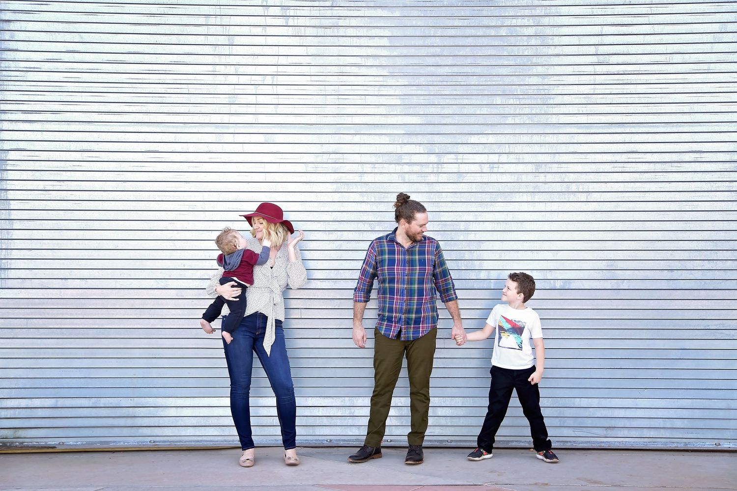 A family of four stands holding hands in front of a white corrugated metal wall, with a mother carrying a baby beside a father and son.