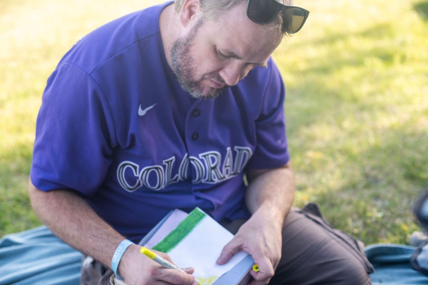 Man in Colorado Rockies jersey writing in notebook while sitting outdoors on grass.