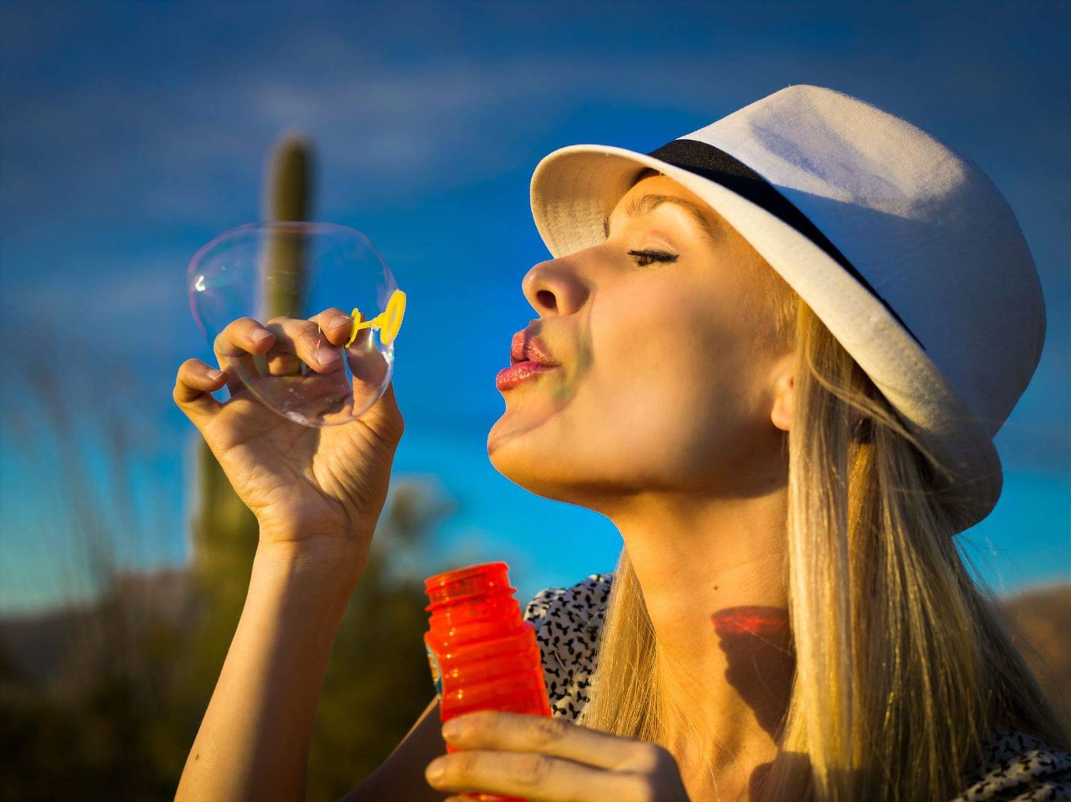 A woman in a white fedora blows a soap bubble from a wand while standing outdoors with a saguaro cactus against a deep blue sky.