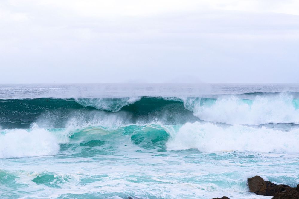 Powerful turquoise ocean waves crashing with white sea foam against rocky shoreline under overcast sky.