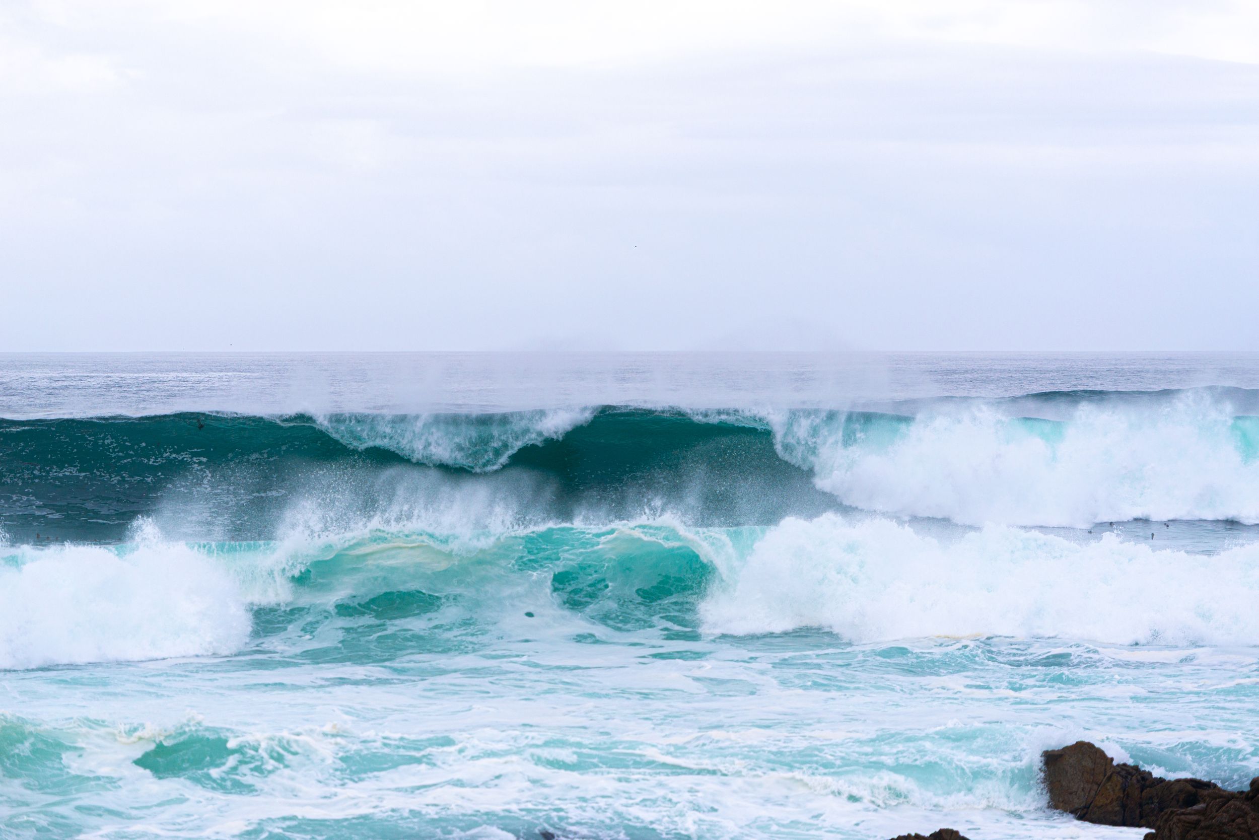 Powerful turquoise ocean waves crashing with white sea foam against rocky shoreline under overcast sky.