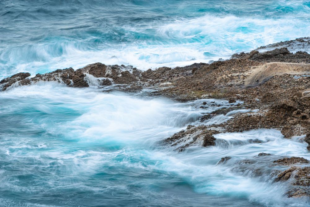 Turquoise ocean waves crashing over rocky coastal shoreline with white sea foam and blue water.