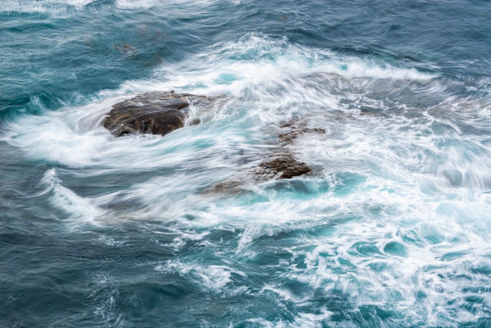 Rocky outcrop surrounded by swirling turquoise ocean waves and white sea foam.