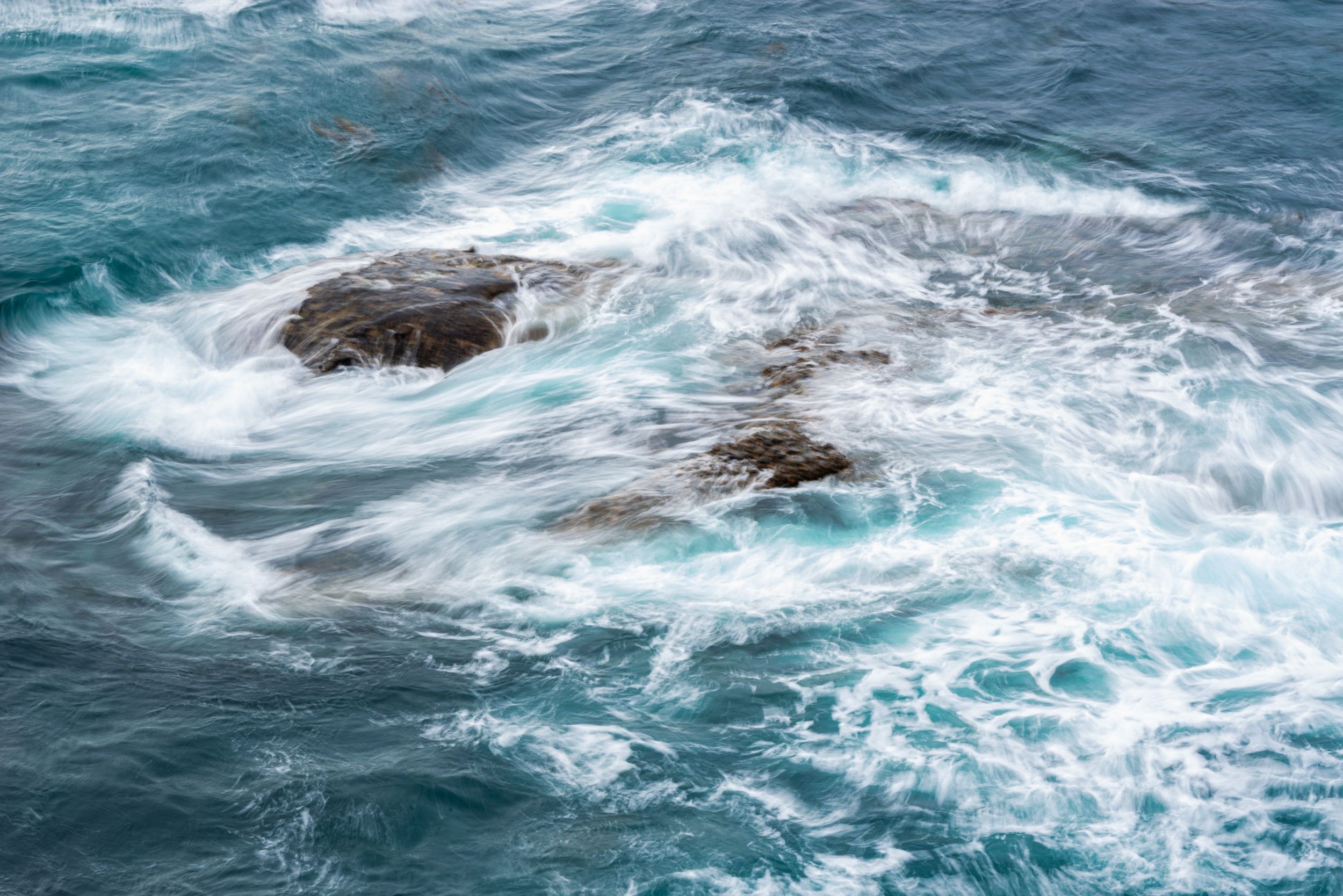 Rocky outcrop surrounded by swirling turquoise ocean waves and white sea foam.