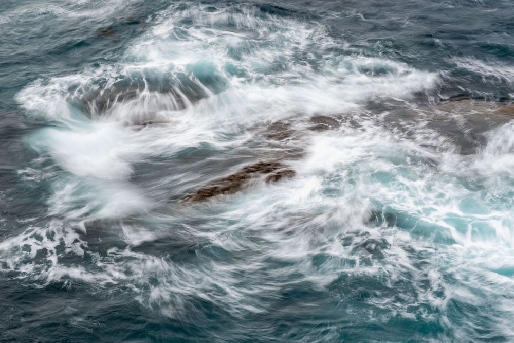 Aerial view of turbulent ocean waves crashing over dark rocks, creating white sea foam in deep blue water.
