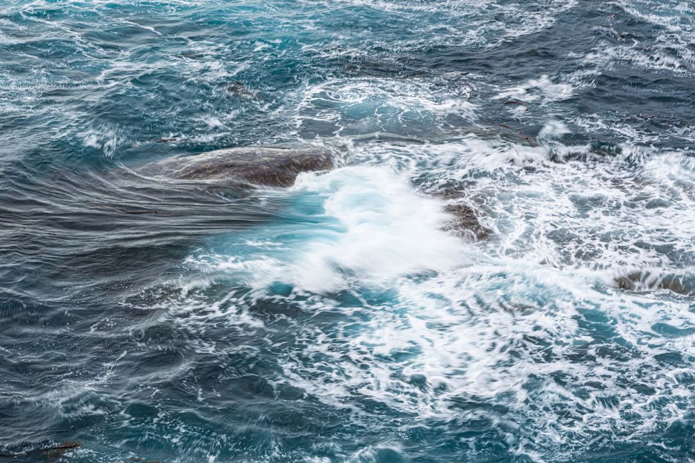 Turbulent ocean waves crashing over dark rocks, creating white sea foam against deep blue-green water.
