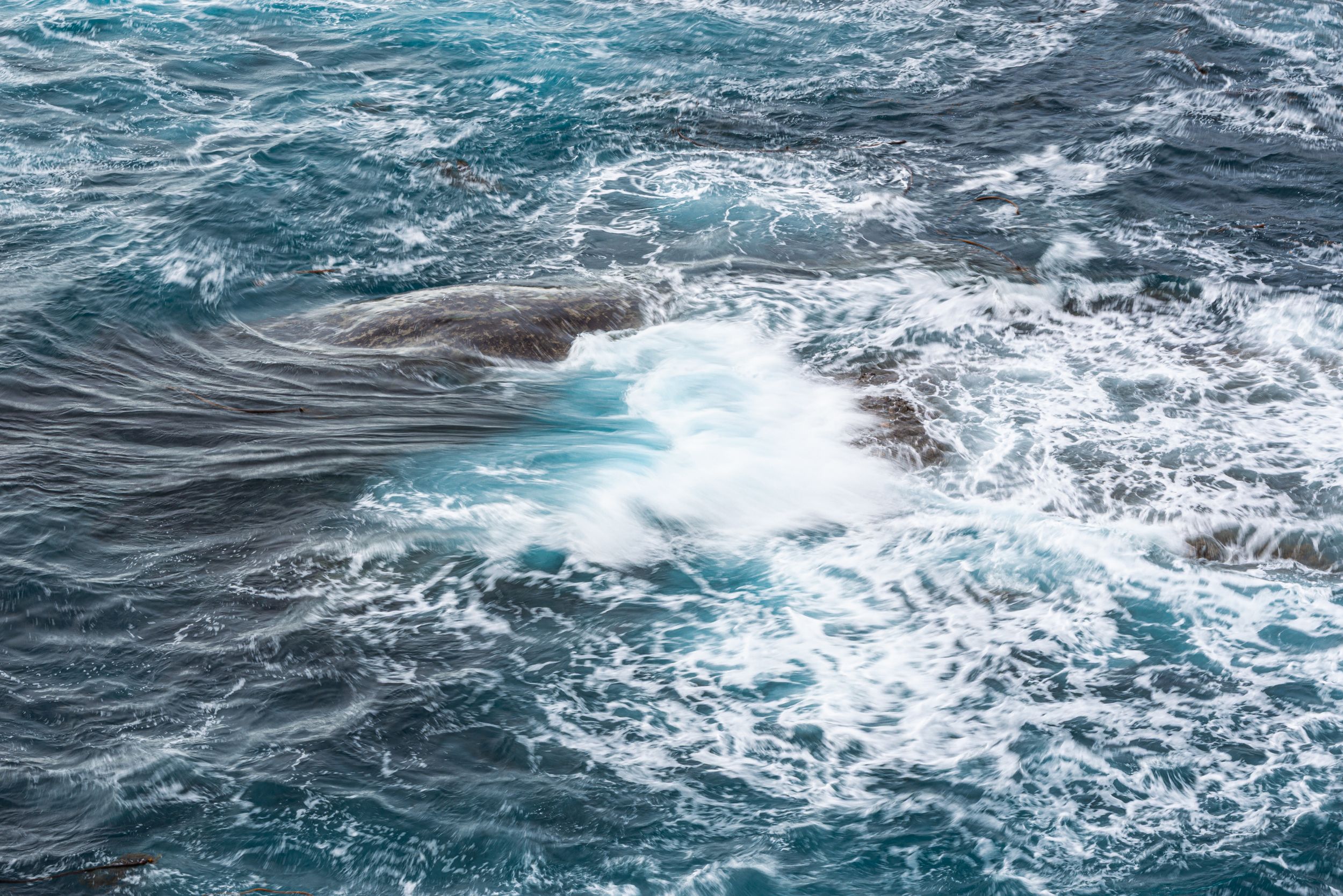 Turbulent ocean waves crashing over dark rocks, creating white sea foam against deep blue-green water.