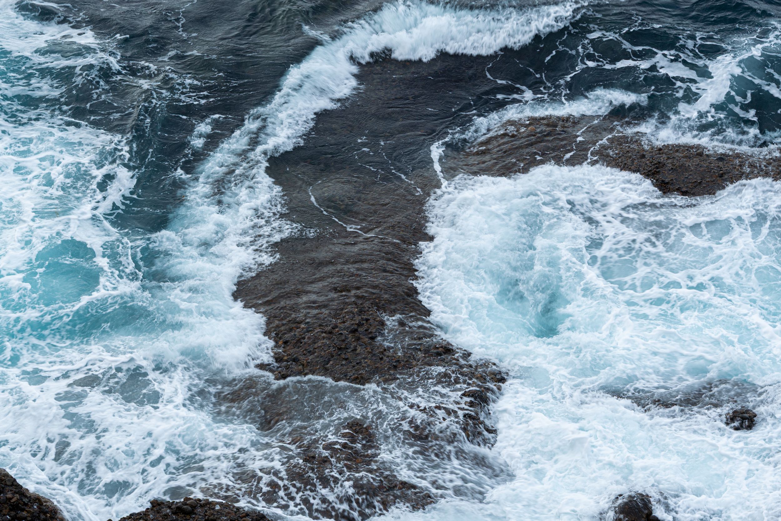 Aerial view of turbulent ocean waves crashing over dark rocky coastline with white sea foam.