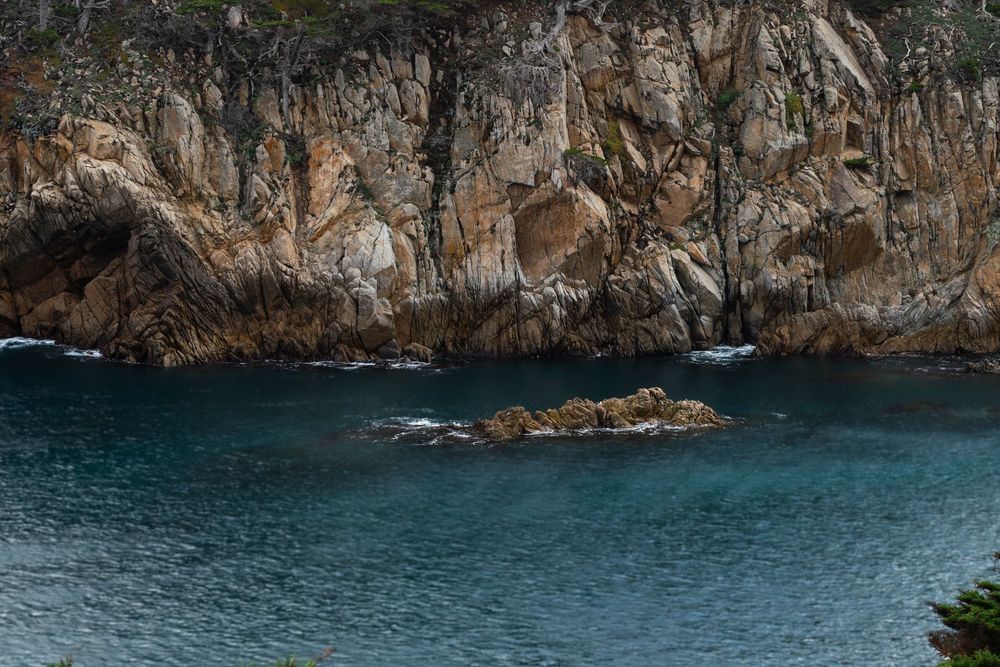 Turquoise coastal waters lap against rugged rocky cliffs with a small rock formation emerging from the calm sea below.