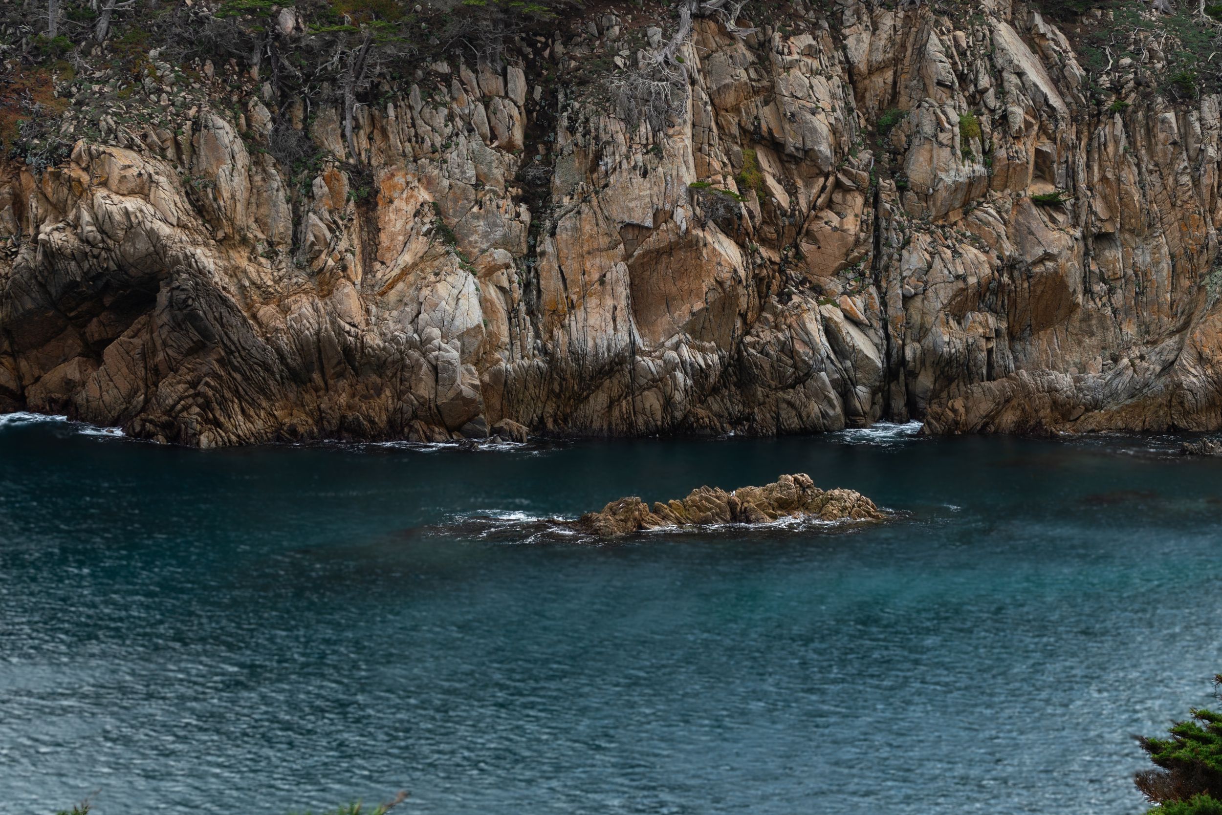 Turquoise coastal waters lap against rugged rocky cliffs with a small rock formation emerging from the calm sea below.