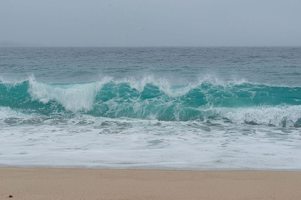 Turquoise ocean waves crashing onto a sandy beach on an overcast day.