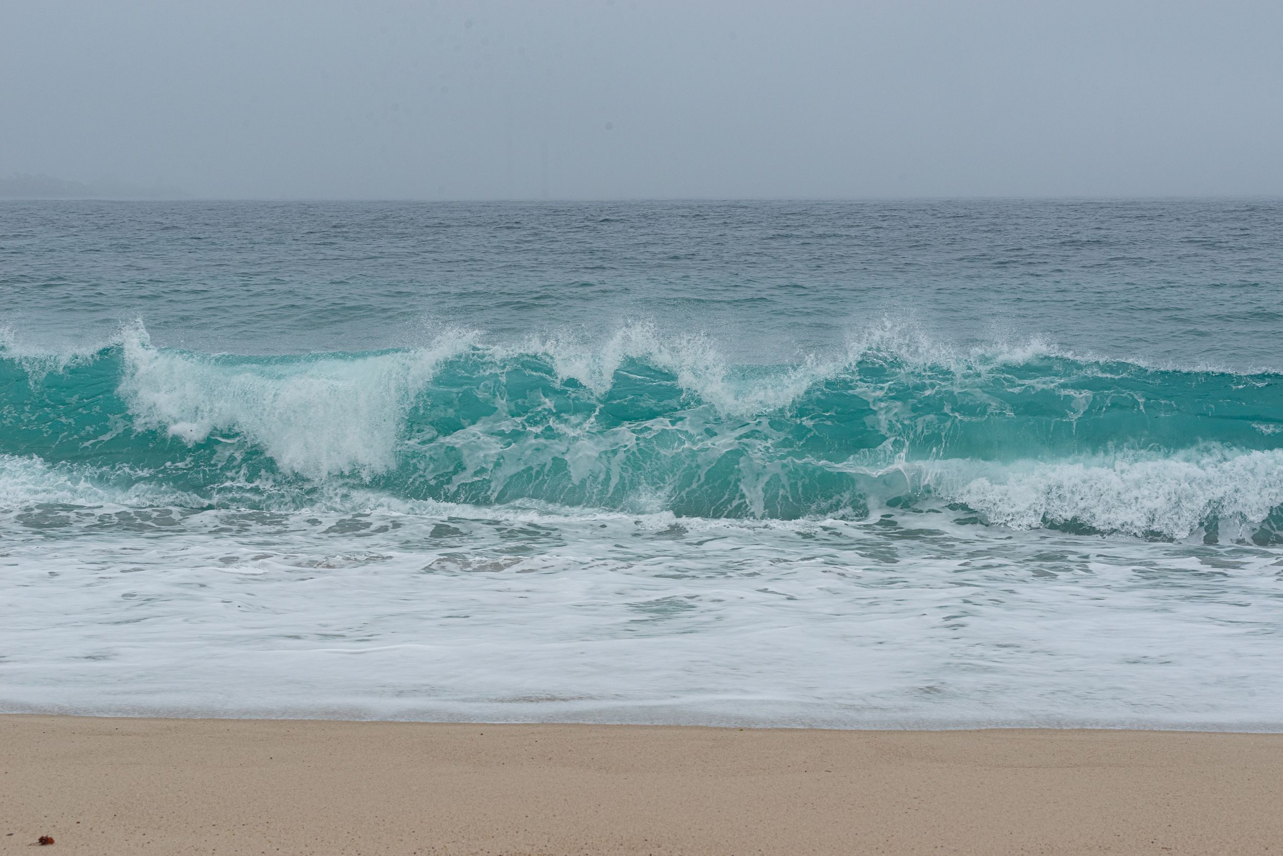 Turquoise ocean waves crashing onto a sandy beach on an overcast day.
