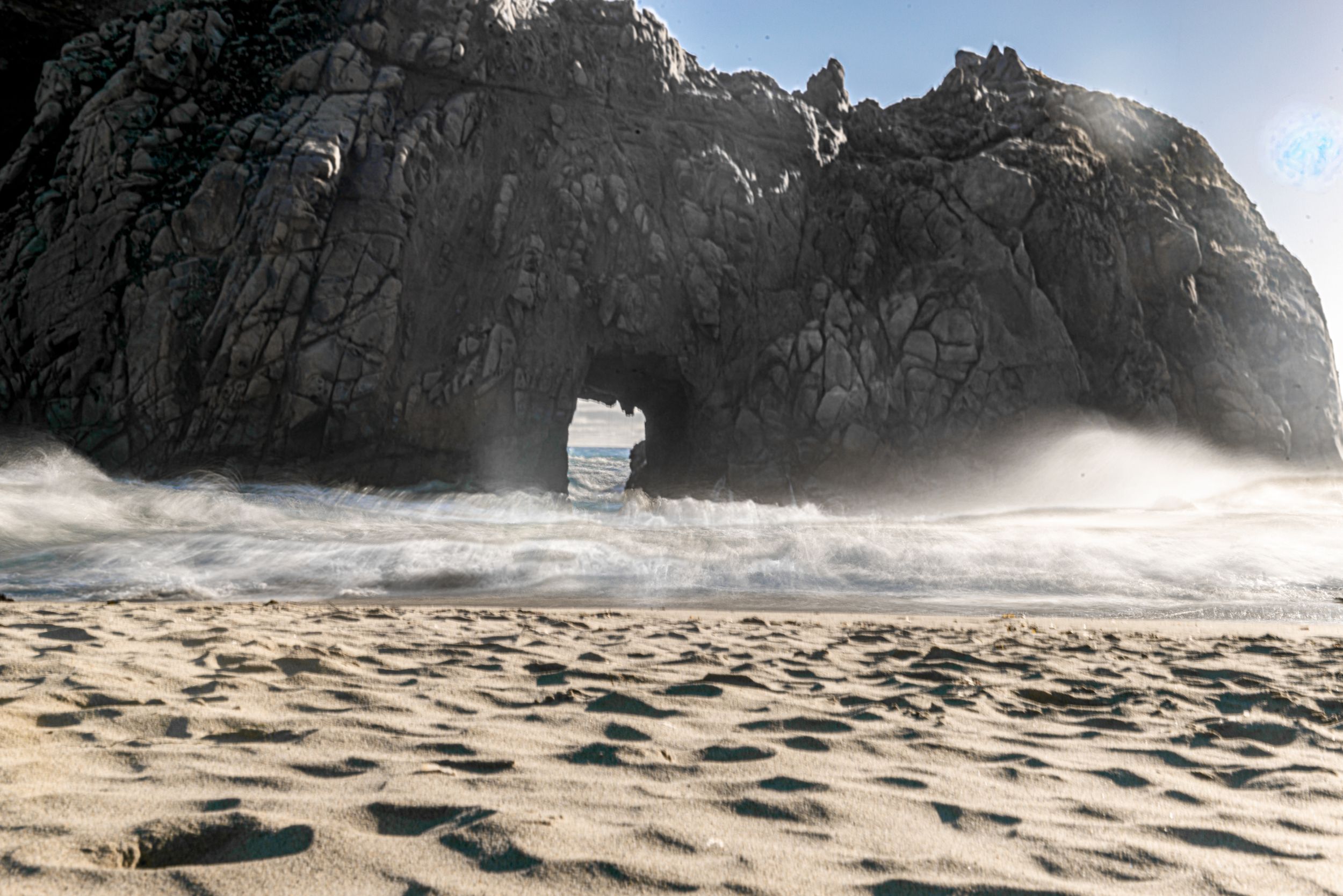 Waves crash through a natural rock arch on a sandy beach, with mist rising against dramatic coastal cliffs.