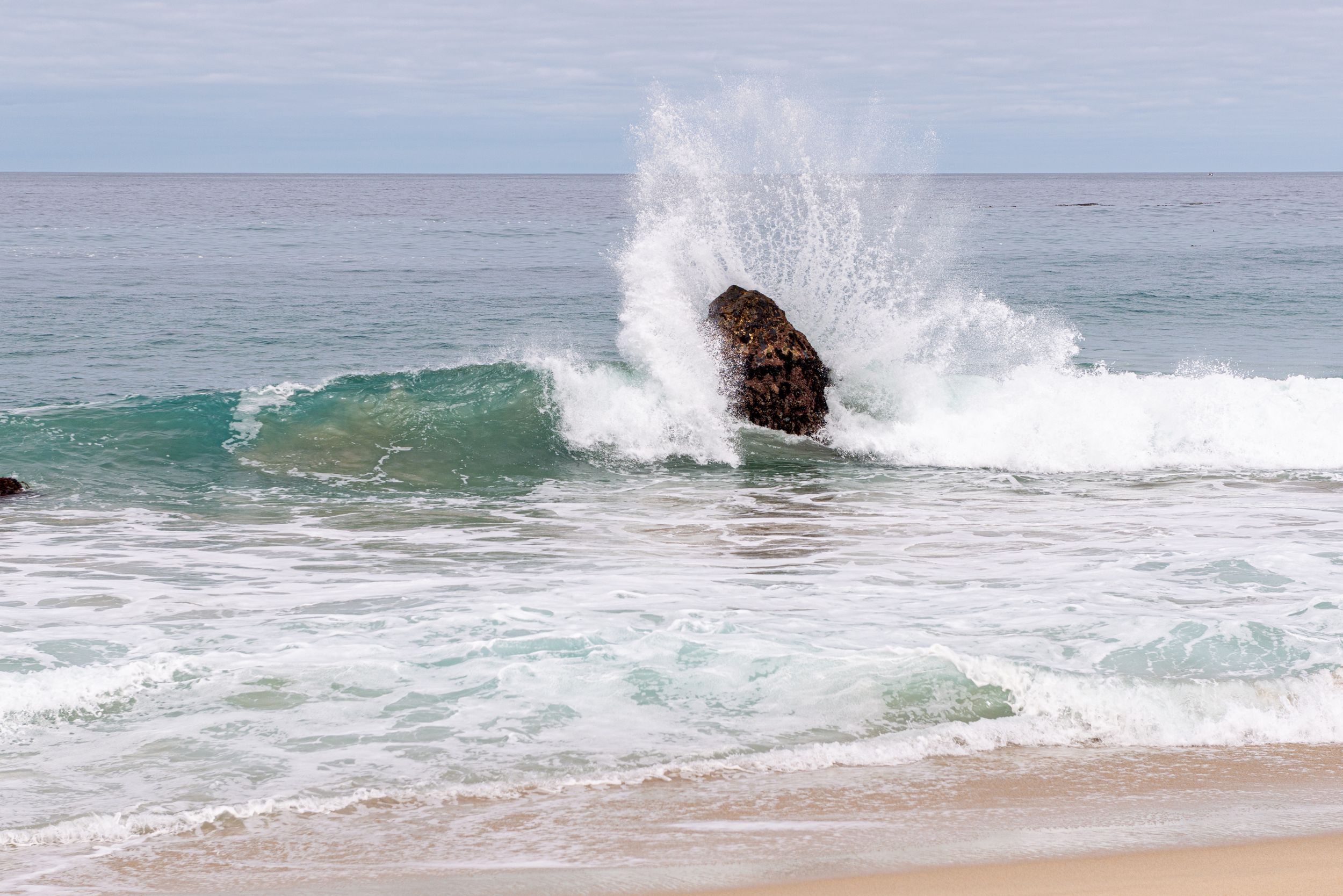Large ocean wave crashing dramatically against a rock on a sandy beach with clear skies.